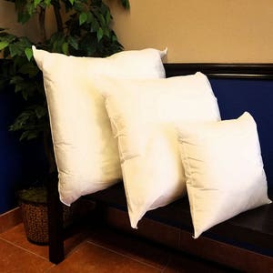 May include: Four white pillows of different sizes are stacked on a dark brown wooden bench. The pillows are made of a soft, white fabric and have a smooth, even surface. The bench is against a wall with a blue bottom half and a tan top half.