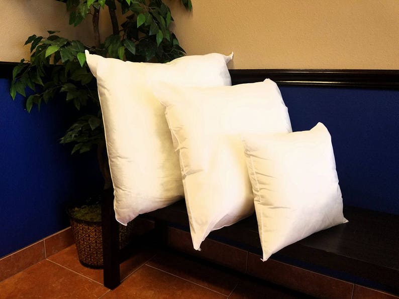 May include: Four white pillows of different sizes are stacked on a dark brown wooden bench. The pillows are made of a soft, white fabric and have a smooth, even surface. The bench is against a wall with a blue bottom half and a tan top half.