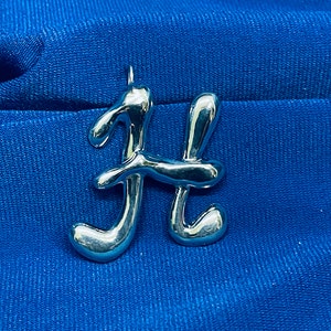 May include: A silver-coloured pendant in the shape of the letter "H" is shown against a blue fabric backdrop. The pendant has a smooth, rounded design and a small loop at the top for a chain.