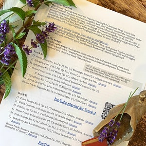 May include: A close-up of a printed sheet of paper with a list of classical music pieces, including composers and their works. The paper is on a wooden table with a small book and a decorative seashell with lavender flowers.