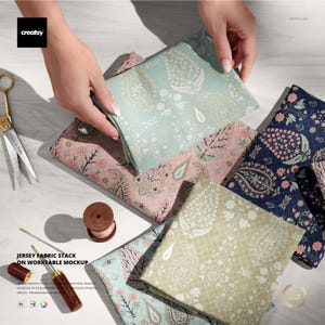 Puede incluir: Muestras de tela de jersey plegadas en varios colores, incluyendo verde claro, rosa y azul marino, cada una con un estampado de cachemira. También se ven tijeras doradas y una bobina de hilo de madera.