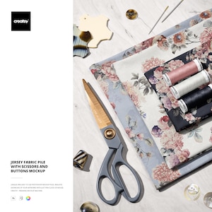 May include: A mockup featuring a jersey fabric pile with floral patterns, scissors, spools of thread, buttons, and a thimble. The scissors have gold blades and gray handles. The fabric has a light blue, white, and dark blue background.