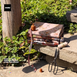 May include: A stack of folded floral fabrics in shades of pink, brown, and cream rests on stone steps. A wooden spool, measuring tape, thimble, and scissors are arranged nearby. The image includes the text "STACK OF FOLDED FABRICS ON GARDEN STONE STEPS MOCKUP."