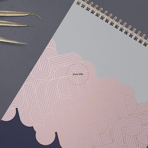May include: A spiral bound notebook with a gray cover and a pink and gold geometric pattern. The pattern is made up of overlapping circles and lines.