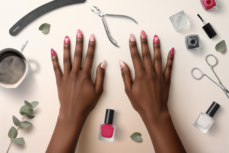 May include: Overhead view of hands with pink and white nail art. Nail polish bottles, nail clippers, scissors, and a coffee cup are arranged around the hands on a light beige surface. A nail file and some greenery are also present.