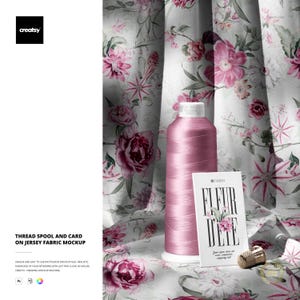 May include: A pink thread spool with a white base and a business card that reads "FLEUR D'ETE" in black text. The card has a floral design. The background is a floral fabric with pink and white flowers.