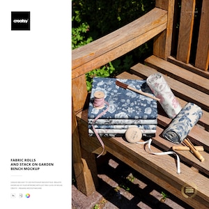 May include: A wooden bench displays fabric rolls and a stack of fabric with a floral pattern in shades of blue and white. A pink measuring tape, wooden tools, and a spool of thread are also present. The text "FABRIC ROLLS AND STACK ON GARDEN BENCH MOCKUP" is visible.