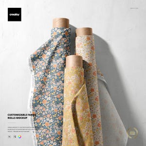 Op de afbeelding: Drie rollen bloemenstof in verschillende kleuren, waaronder blauw, geel en crème. De stof is opgerold rond kartonnen kokers. De afbeelding bevat de tekst "CUSTOMIZABLE FABRIC ROLLS MOCKUP".