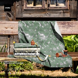 May include: A stack of green fabric samples with a white and gray bird and floral pattern, displayed on a wooden bench. A wooden spool, a measuring tape, and a small wooden bottle are also present. The text "FABRICS ON RUSTIC FARMHOUSE BENCH MOCKUP" is visible.