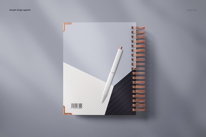 Op de afbeelding: Een wit spiraalnotitieblok met een geometrisch ontwerp in zwart, wit en grijs. Het notitieblok heeft een koperkleurige binding en een witte pen ligt erop.
