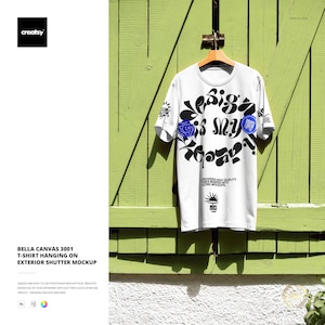 May include: A white Bella Canvas 3001 t-shirt with a graphic design, hanging on a wooden hanger. The design includes black and blue floral elements and the text "Design is my Utopia". The shirt is displayed against a green wooden shutter.