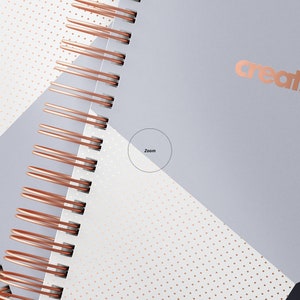 Op de afbeelding: Een close-up van een spiraalgebonden notitieboek met een ros&eacute;gouden draadbinding. De omslag is een geometrisch ontwerp met witte, grijze en zwarte secties. Het woord "creatsy" is in ros&eacute;goud gedrukt op de grijze sectie.