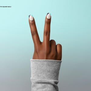 May include: A hand with dark skin showing the peace sign, index and middle fingers raised. The square nails have a white base, navy blue tips, and a light blue accent. The wrist is covered by a grey ribbed sleeve. The background is a gradient.