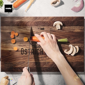 May include: A rectangular end-grain wood cutting board with the text "OSTRICH ARIZONA" and "HONEST" printed on it. A person is cutting a carrot with a knife on the board. Carrots and mushrooms are scattered around the board.