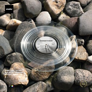 Peut inclure: Un disque vinyle transparent avec le mot "TOGETHER" imprimé sur l'étiquette, posé sur un lit de pierres grises et brunes. Les rainures du disque sont visibles, et l'image comprend le texte "CLEAR VINYL RECORD PLACED ON THE ROCKS MOCKUP".