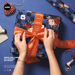 Könnte beinhalten: Ein Weihnachtsgeschenk, verpackt in Papier mit floralem und kariertem Muster, wird mit einem orangefarbenen Band gebunden. Auf dem Band steht "Merry Christmas". Das Geschenk liegt auf einer blauen Oberfläche.
