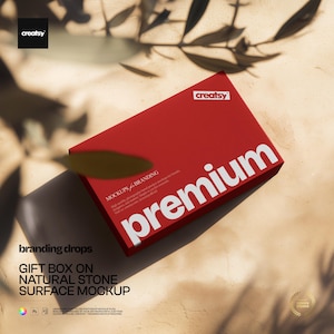 May include: A red gift box with the word "premium" in large white letters. The box is on a natural stone surface with shadows and the text "branding drops" and "GIFT BOX ON NATURAL STONE SURFACE MOCKUP". The image includes the "creatsy" logo.