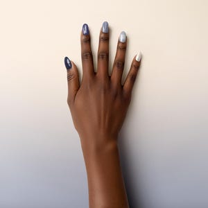 May include: A hand with long, oval nails painted in shades of blue, grey, and white. The nails feature solid colours and a glittery finish. The hand is set against a cream and light blue gradient background.