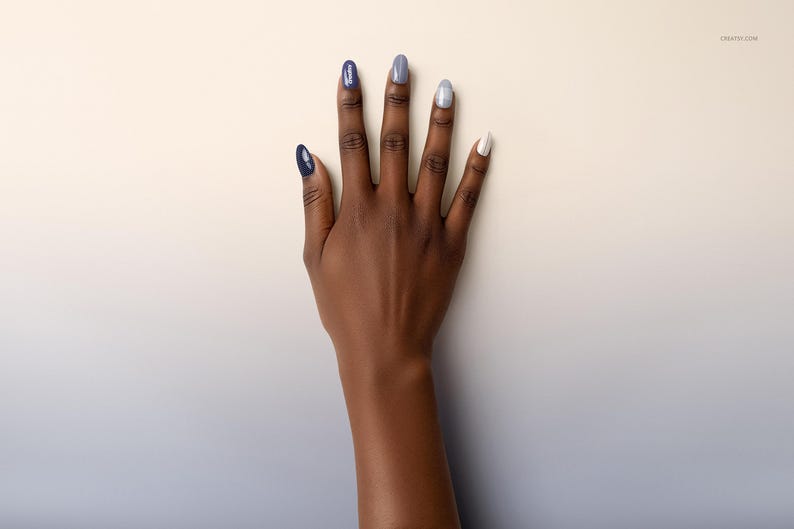 May include: A hand with long, oval nails painted in shades of blue, grey, and white. The nails feature solid colours and a glittery finish. The hand is set against a cream and light blue gradient background.