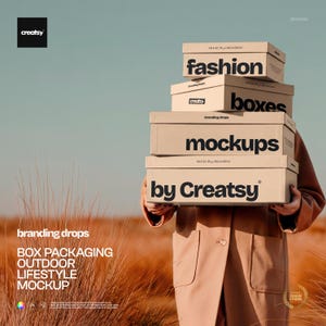 May include: A person holding a stack of beige boxes with the words "fashion", "boxes", "mockups", and "by Creatsy" printed in black. The boxes are in an outdoor setting with tall, dry grass and a blue sky.