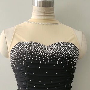 Peut inclure: Une robe de patinage artistique noire avec un d&eacute;collet&eacute; en mesh transparent et un corsage recouvert de strass. La jupe est superpos&eacute;e et a un ourlet festonn&eacute;.