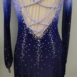 May include: A long-sleeved figure skating dress with a lavender and blue ombre design. The dress has a criss-cross back with a keyhole opening and is embellished with rhinestones.
