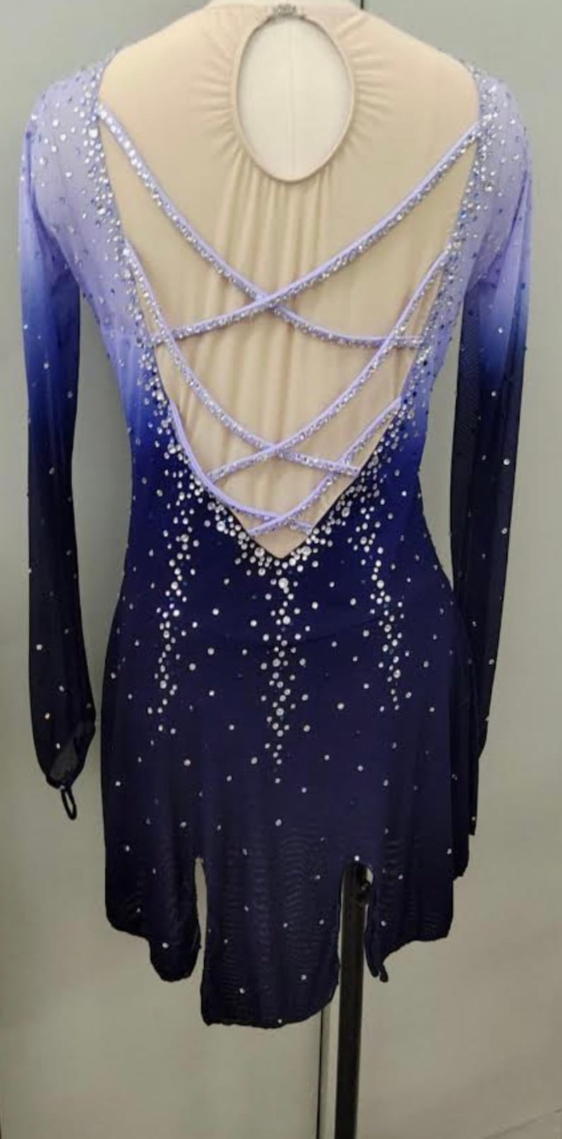May include: A long-sleeved figure skating dress with a lavender and blue ombre design. The dress has a criss-cross back with a keyhole opening and is embellished with rhinestones.