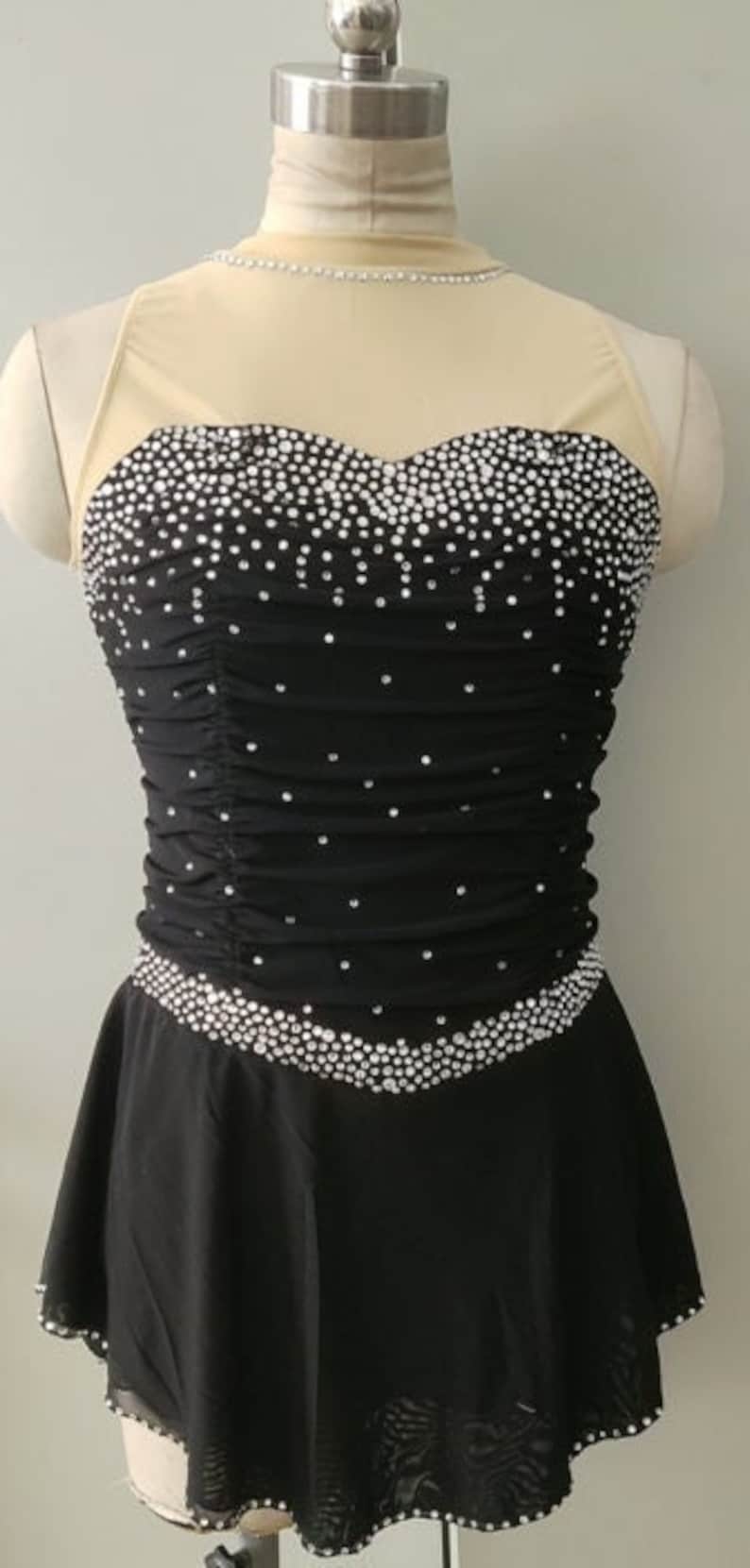 Peut inclure: Une robe de patinage artistique noire avec un d&eacute;collet&eacute; en mesh transparent et un corsage recouvert de strass. La jupe est superpos&eacute;e et a un ourlet festonn&eacute;.