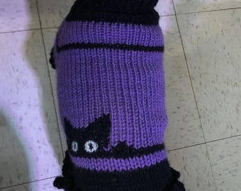 May include: A purple and black knitted dog sweater with a black cat design. The sweater has a ruffled hem.