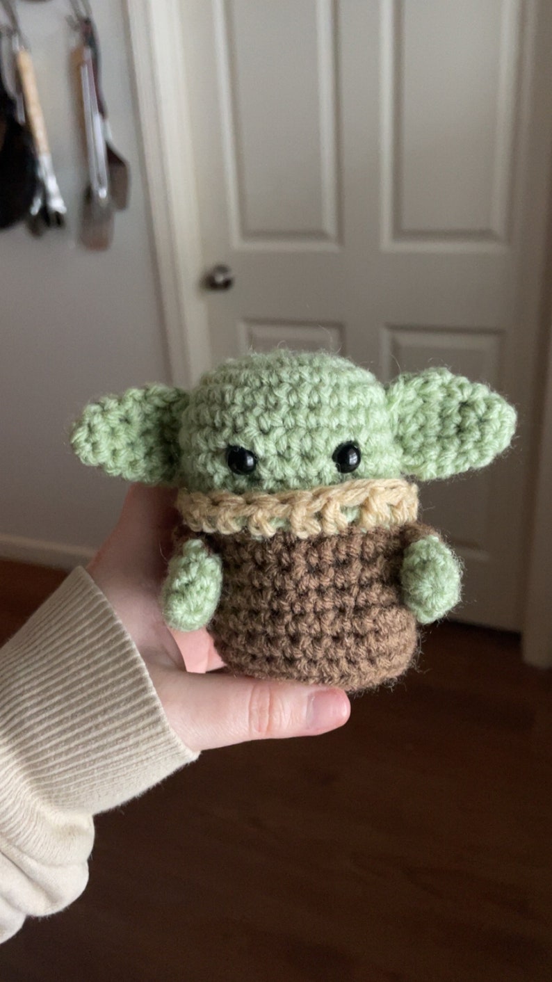 May include: A crocheted green and brown Baby Yoda plush toy with large ears and black eyes. The toy is being held in a person's hand.