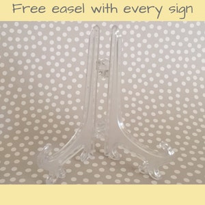 May include: A clear plastic easel stand for displaying signs or artwork. The stand has a simple design with three legs and a flat top.