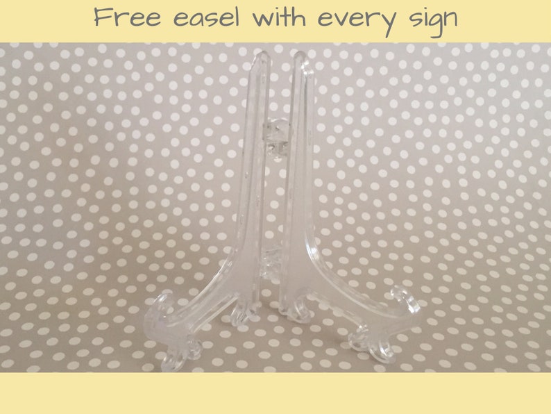 May include: A clear plastic easel stand for displaying signs or artwork. The stand has a simple design with three legs and a flat top.