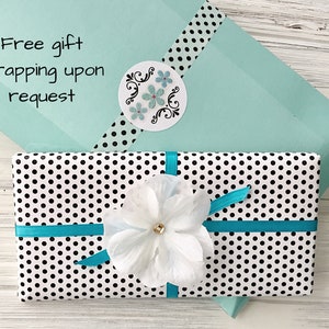 May include: A white gift box with black polka dots wrapped with a turquoise ribbon and a white flower. The box is sitting on a light wood surface. The text "Free gift wrapping upon request" is visible on a light blue envelope.
