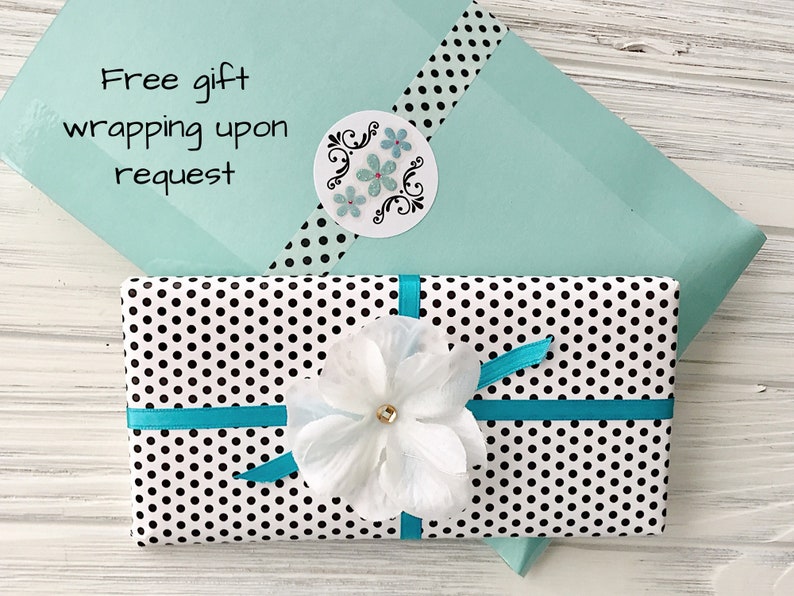 May include: A white gift box with black polka dots wrapped with a turquoise ribbon and a white flower. The box is sitting on a light wood surface. The text "Free gift wrapping upon request" is visible on a light blue envelope.