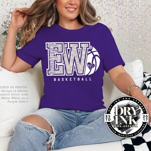 a model sitting on the sofa wearing a bella and canvas 3001 tshirt in the color purple with a custom glitter basketball design in a two letter version of initials with a basketball that is personalizable. The tshirt graphic is in the color glitter confetti silver and matte white.