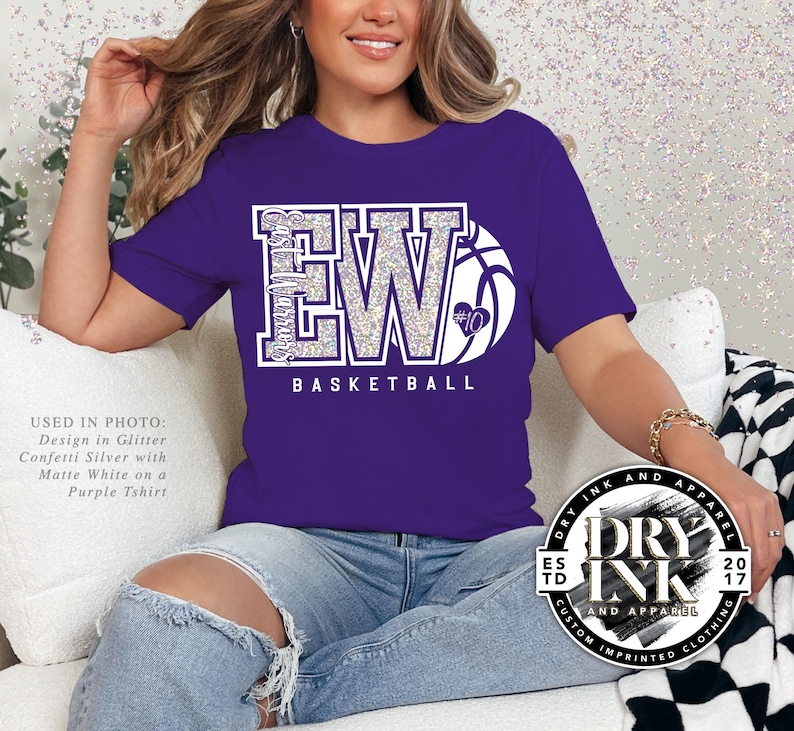 a model sitting on the sofa wearing a bella and canvas 3001 tshirt in the color purple with a custom glitter basketball design in a two letter version of initials with a basketball that is personalizable. The tshirt graphic is in the color glitter confetti silver and matte white.
