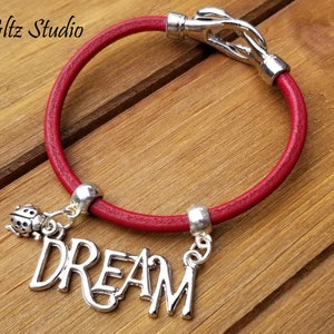 Könnte beinhalten: Rotes Lederarmband mit einem silbernen Verschluss und zwei Anhängern. Ein Anhänger ist eine silberne Marienkäfer und der andere ist ein silberner Anhänger mit dem Wort "DREAM" in Kursivschrift.