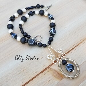 May include: Black and white beaded necklace with a silver wire pendant. The pendant is shaped like a teardrop and has a black and white bead at the bottom. The necklace is on a wooden surface.