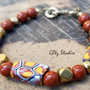 Könnte beinhalten: Ein rotes und goldenes Perlenarmband mit einer geometrisch gemusterten Perle. Das Armband hat einen Verschluss und ist aus Natursteinen und Metallperlen gefertigt.