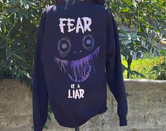Könnte beinhalten: Schwarzer Kapuzenpullover mit einem lila und weißen Grafikdruck eines lächelnden Gesichts mit dem Text "FEAR IS A LIAR" auf der Rückseite.