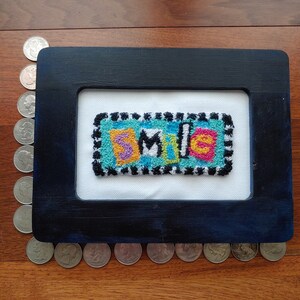 May include: A framed piece of needlepoint art with the word "SMILE" spelled out in colorful yarn. The frame is black with a blue inner border.