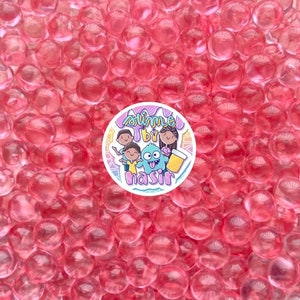 May include: A close-up of a collection of translucent, pink orbeez. A circular sticker in the center reads "Slime by Nasir" with cartoon illustrations of people and a monster.