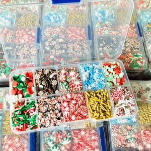 May include: A collection of clear plastic containers filled with various Christmas-themed sprinkles. The sprinkles include red, white, and green candies in shapes like snowflakes, candy canes, and round shapes. The containers are arranged in rows.