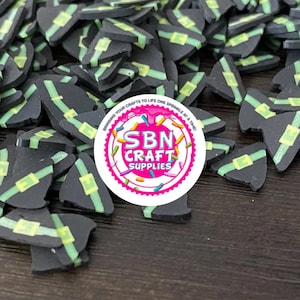 May include: A large pile of black witch hat shaped polymer clay charms with green and yellow accents. The charms are scattered on a dark wood surface. A sticker with the text "SBN CRAFT SUPPLIES" is in the center of the image.