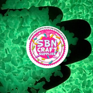 Puede incluir: Primer plano de una gran cantidad de brillantina verde que brilla en la oscuridad. La brillantina está en una mano, y un logotipo rosa y blanco con el texto "SBN Craft Supplies" es visible en el fondo.
