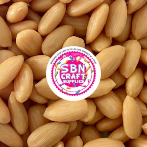May include: Close-up of a pile of blanched almonds, showcasing their smooth, pale beige color and elongated shape. The image includes a circular logo with the text "SBN CRAFT SUPPLIES".