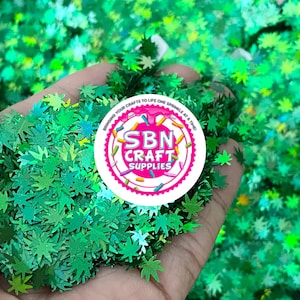 May include: A close-up of a hand holding a pile of green glitter shaped like maple leaves. The glitter is iridescent and has a holographic finish. The background is a pile of the same glitter. A sticker with the text "SBN CRAFT SUPPLIES" is visible in the image.