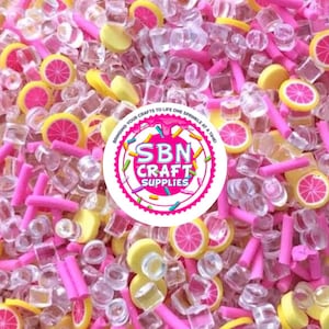 May include: A vibrant mix of craft supplies featuring pink, yellow, and clear elements. The assortment includes pink cylinders, yellow and pink citrus slices, and clear cube-shaped pieces. A circular logo with the text "SBN CRAFT SUPPLIES" is also visible.