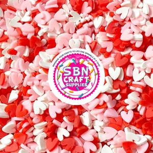 May include: A close-up shot of a mix of heart-shaped sprinkles in shades of red, pink, and white. The sprinkles are scattered, creating a textured surface. A circular logo with text is visible in the image.
