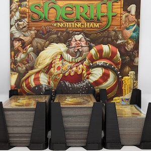 Pode incluir: Um organizador de jogos de cartas preto com três compartimentos, cada um com um monte de cartas. O organizador está na frente de uma caixa de jogo de tabuleiro com uma ilustração de desenho animado de um xerife com o texto "Sheriff of Nottingham".